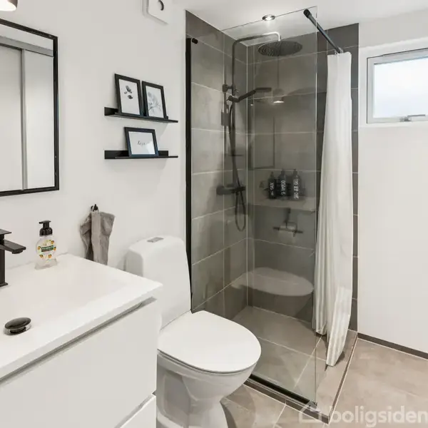 Badeværelse med en hvid vaskenhed, spejl og sorte armaturer. Bruseniche med glasvæg og fliser. Hylder med billeder over toilettet. Lille vindue giver naturligt lysindfald.