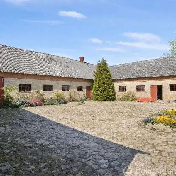 Gårdsplads med brostensbelægning, omgivet af to store, langstrakte staldbygninger med røde døre og vinduer. En høj busk står centralt, og blomsterbede kantet gårdspladsen.