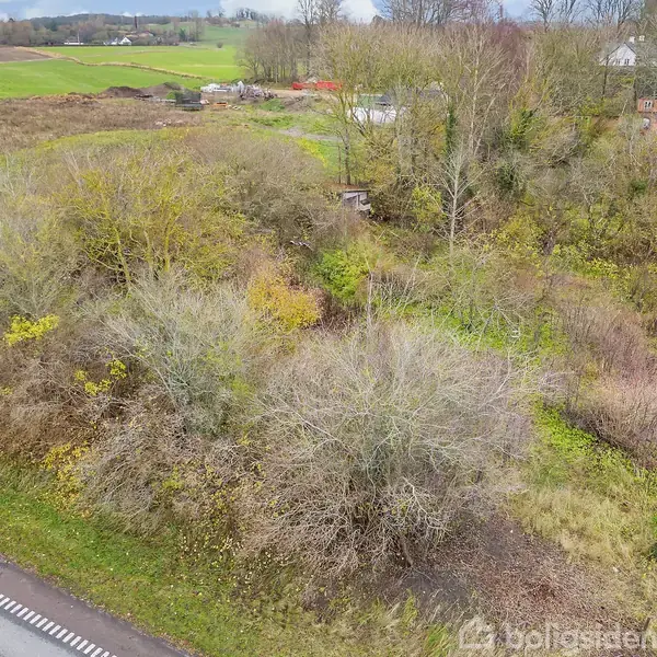 En buskbevokset grund står ubenyttet ved siden af en asfaltvej. Skilte omgivet af marker og spredte boligbyggerier kan ses i baggrunden. Området er naturskønt og frodig.