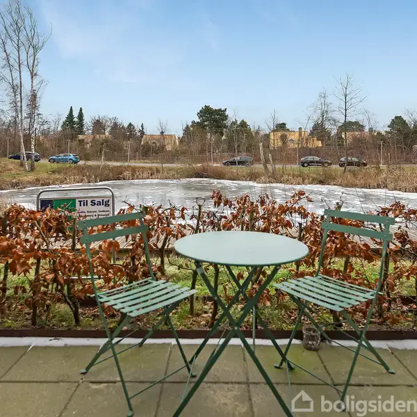 Et grønt cafésæt står på en stenbelagt terrasse foran en hæk med en lille, frossen sø i baggrunden. Et skilt med teksten "Til Salg" er synligt.