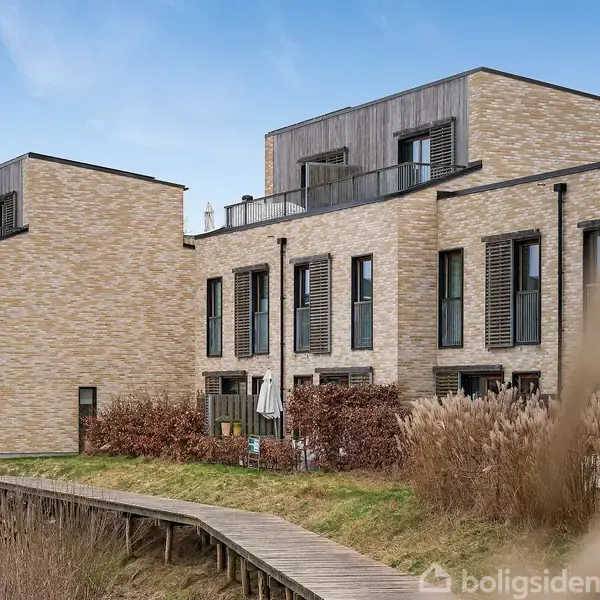Rækkehuse med flade tage og store vinduer står stille langs en gangbro i et grønt boligområde. Murværket er i lysebrune nuancer, og planter vokser nær facaden.