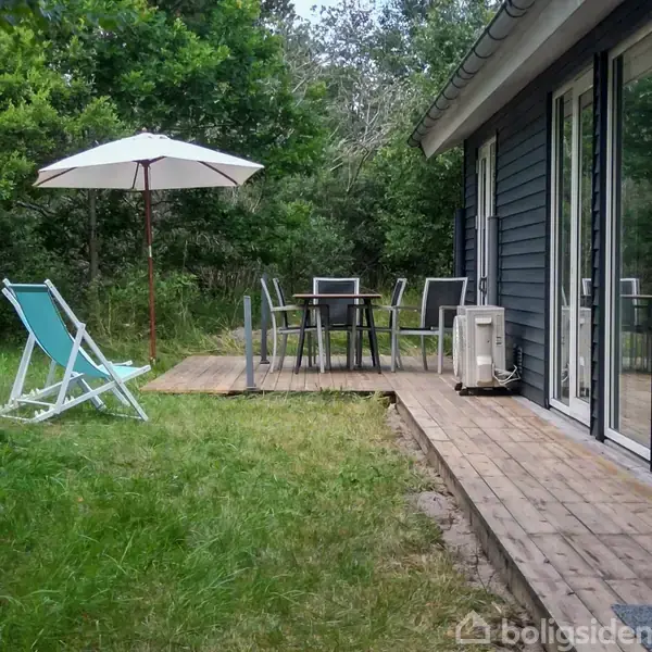 En terrasse har et bord med fire stole under en parasol. Ved siden af er der to strandstole på græsplænen. Til højre ses et hus med store glaspartier.
