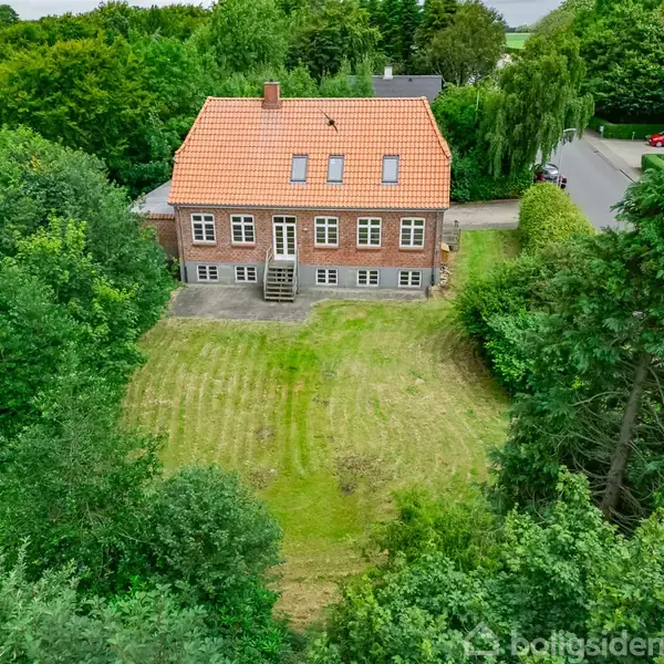 Hus i mursten med rødt tegltag og seks vinduer, omgivet af grøn have. Det ligger for enden af en klippet græsplæne, omkranset af tætte træer og buske.