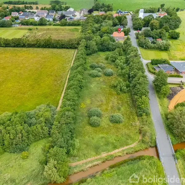 En lang, smal mark med træer løber parallelt med en vej. Omkringliggende grønne områder og huse ligger i det fjerne, skaber en landlig atmosfære.