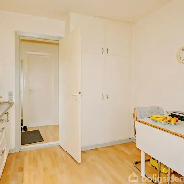 En lys køkken med trælaminatgulv, hvor en kaffemaskine og mikroovn står på en bordplade. Et bord ved væggen har frugt på. Et vægur viser klokken 10 over 10.