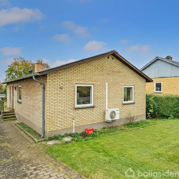 Et gult murstenshus med en græsplæne og indkørsel ved siden af. En grøn hæk omkranser haven, og en blå himmel med skyer ses i baggrunden.