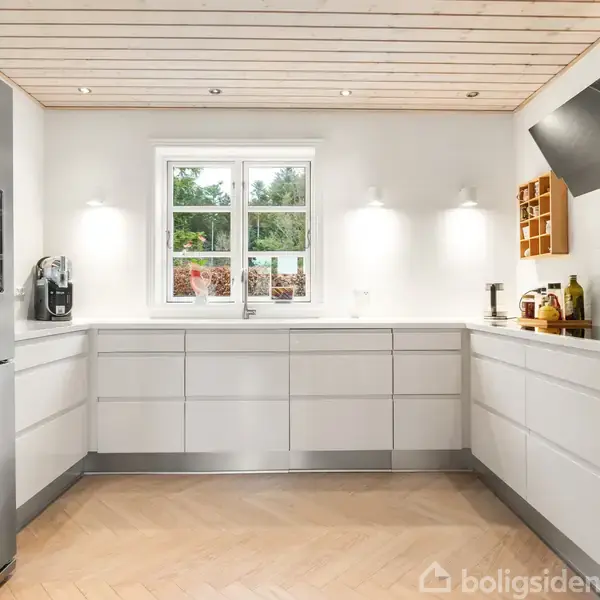 Et moderne køkken med hvide skabe, stort køleskab og indbyggede ovne. En kaffemaskine står på bordpladen. Rummet har trægulv og et vindue, som giver udsigt til en have.