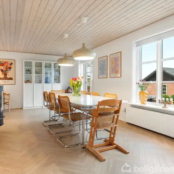 Et spisestuebord med træstole står i midten af et lyst rum. To lamper hænger over bordet med blomster i vase. Store vinduer viser en udsigt til et hus.