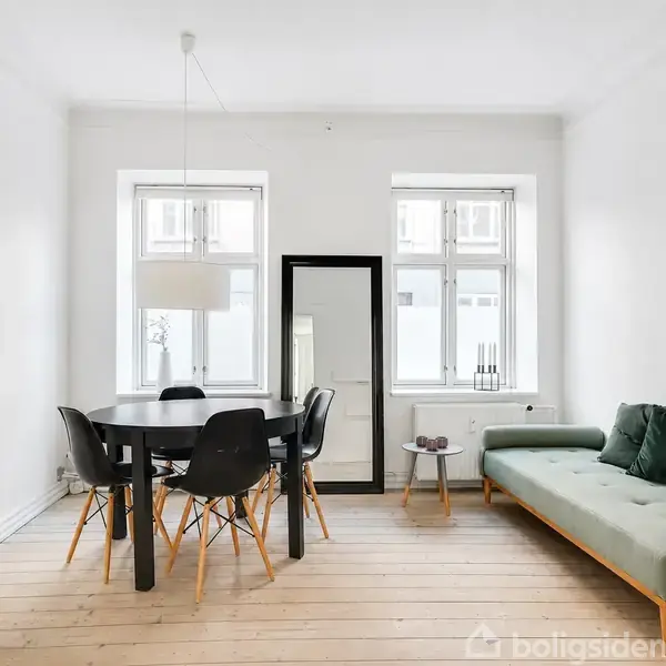 Et lyst rum med et rundt spisebord og fire sorte stole står på et trægulv. Bagved er en grøn sofa, et stort spejl, og to vinduer, der lader lyset strømme ind.