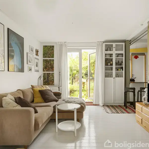 Sofa med puder står i stue, omkranset af kunst på væggene. To runde borde foran sofaen. Døre åbner til have med grøn udsigt. Tv står til højre.