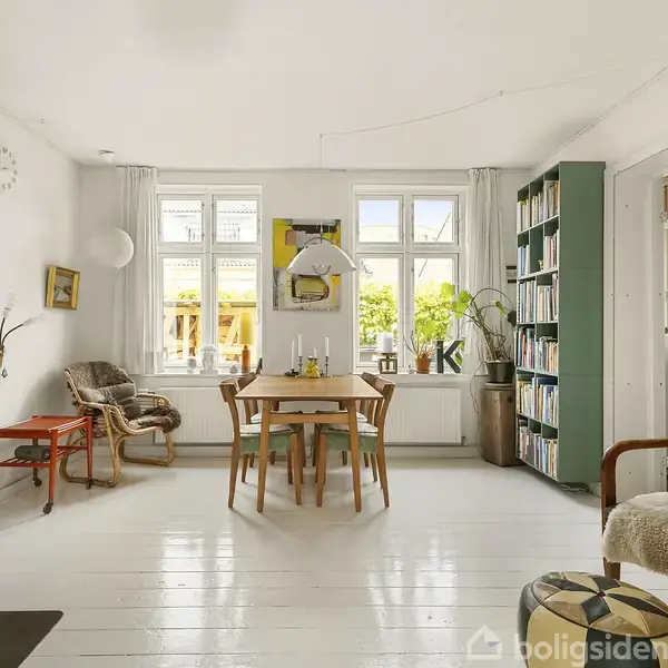 Et lyst rum med en brændeovn til venstre, pejsen brænder. Et spisebord med fire stole står midt i rummet. Bogreol og sofa er placeret til højre. Store vinduer giver udsigt til en grøn have.