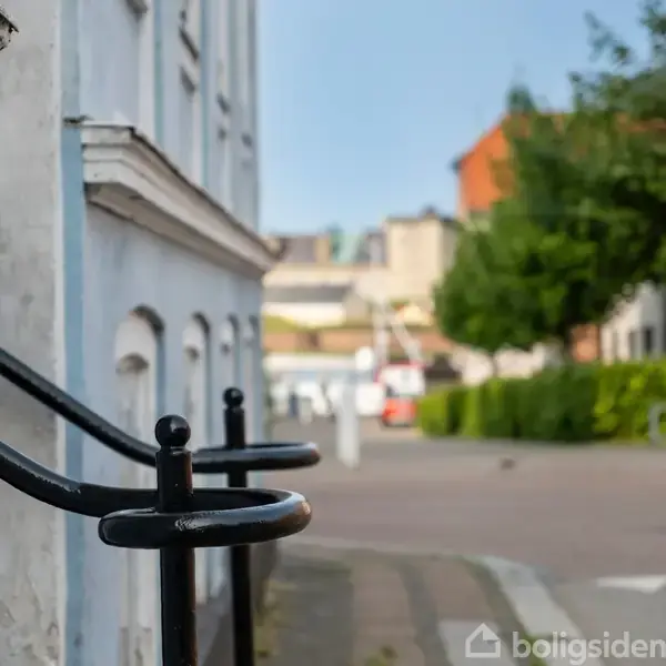 Hvide byhus-rækværk løber langs sidden af bygning, i en stille gade med træer og bygninger i baggrunden.