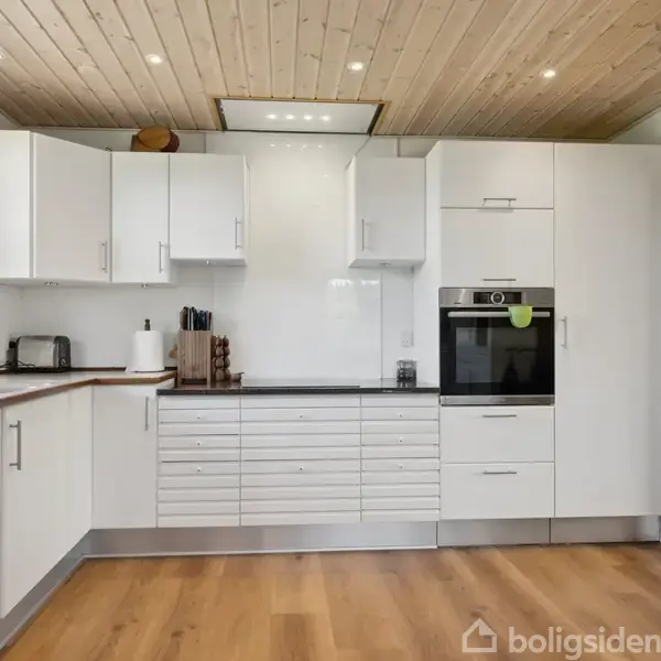 Hvidt modernistisk køkken med hvilende overskabe og indbygget ovn; ligger i et rum med trægulv og træbeklædt loft, ved siden af et stort vindue der giver naturligt lys.