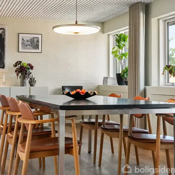 Spisebordet med frugtskål i træstole-stil står i moderne spisestue. Væggene har kunstværker, et skab med lampe, og store vinduer med planter giver naturligt lys.