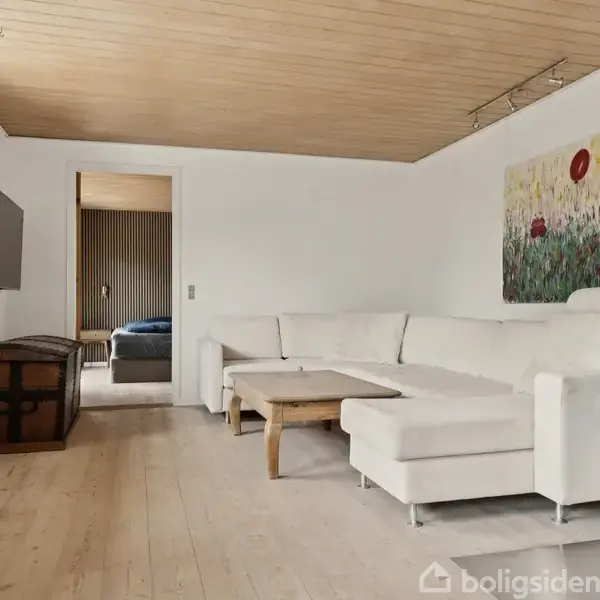 Hvid hjørnesofa står i en stue med træloft, fladskærms-tv på væggen, trækommode og pejs. Dør fører til soveværelse med blåt sengetæppe. Maleri hænger over sofaen.