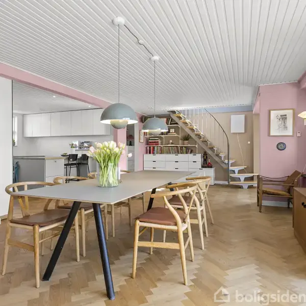 Moderne spisestue med lange spisebord omgivet af træstole. Over bordet hænger en lampe. Baggrunden inkluderer et køkkenområde, en trappeopgang og lyserøde vægge med kunst. Enkommode står ved siden af med bøger og blomster.
