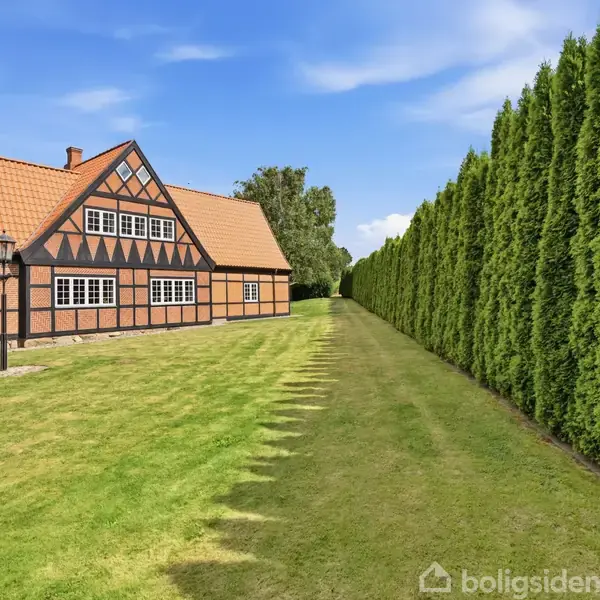 Hus med korsvirkestil står på en velplejet græsplæne ved siden af en række høje, tætte buske. Skyfri blå himmel og dagslys fremhæver landskabet.