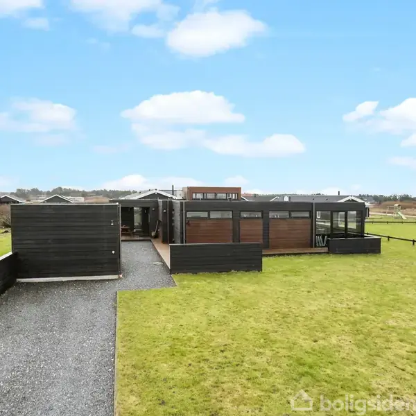 Et moderne hus med sort træfacade står midt i en stor, grøn græsplæne. Grusstien fører til hovedindgangen, omgivet af hegn og andre huse i det fjerne.