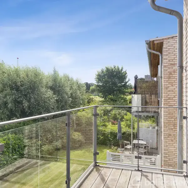 En balkon med glasrækværk vender ud mod en grøn have og frodig bevoksning. Bygningen er af mursten, og sollys skinner ind. Himmel er klar med få skyer.
