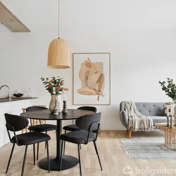 Spisebord med sorte stole står ved siden af en grå sofa og et rundt bord. Kunstværk hænger på væggen i et moderne, lyst rum med trægulv og åben køkkenadgang.