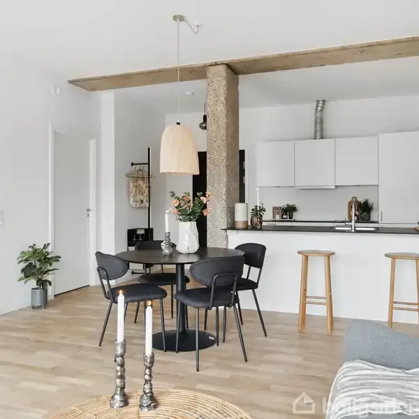 En stue med træbord og sorte stole står foran et moderne, åbent køkken med hvide skabe. Rummets indretning er minimalistisk, og der er planter samt en stor pendellampe.