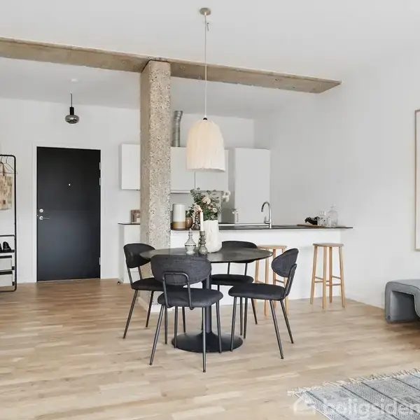 Sort rundt spisebord med fire stole står i moderne stue. Køkken med hvidt interiør og barkrakker i baggrund. Stor plante og abstrakt kunstværk pryder væggen.