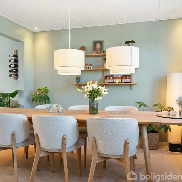 Et spisebord med seks hvide stole står i et lyst rum. To pendellamper hænger over bordet, der er dekoreret med blomster. Grønne planter og en hylde pynter væggene.