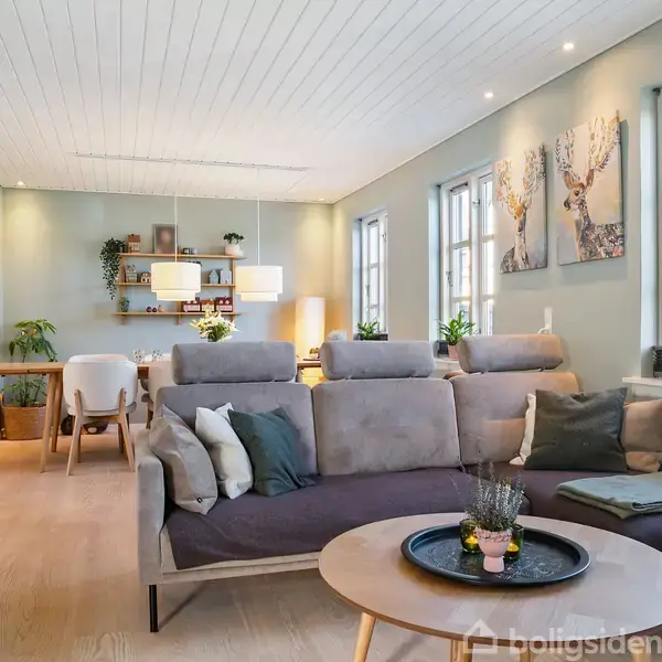 Stue med grå sofa, sofabord med bakke, og vægkunst i lyse nuancer. Spisebord og stole i baggrunden. Store vinduer og planter bringer naturligt lys og grønne elementer ind.