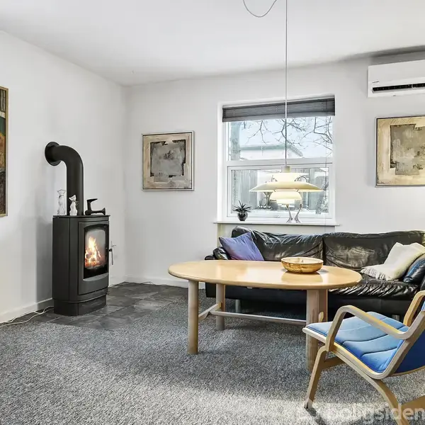 Stue med brændeovn, tændt og afgiver varme. En sort sofa med farverige puder står bag et rundt træbord. To lamper lyser rummet, som har kunst på væggene.