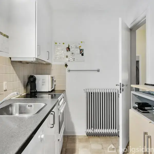 Køkken med sort bordplade og vask, kaffemaskine og mikrobølgeovn. På væggen ses et radiator under opslagstavle. Til højre er en åben dør ind til en gang.