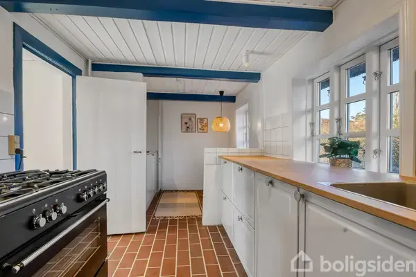 Komfur står i køkkenet, ved en væg, omgivet af hvidmalede skabe, træbordplade og store vinduer. Loftet har blå bjælker, og gulvet er belagt med røde klinker, hvilket skaber en lys atmosfære.