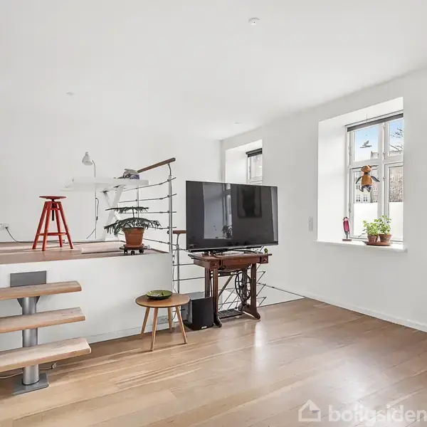 Et moderne rum med trægulv og hvide vægge. Et tv står på et bord, planter er i vinduet og på gulvet. En plakat hænger på væggen. Trapper fører op til et forhøjet område med stol og planter.