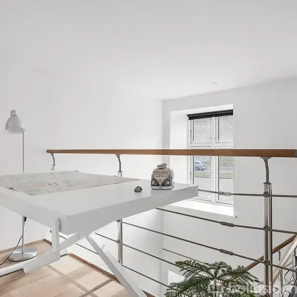 Tegnebord med lampe og genstande står ved en trappeafsats i et lyst rum med vinduer. En rød skammel står ved siden af bordet, og en plante er placeret nær trappen.