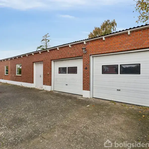 Rød murstensbygning med to hvide garageporte og en dør står i et udendørs område. Træer og en asfaltvej omgiver bygningen, der ser funktionel ud til opbevaring eller parkering.