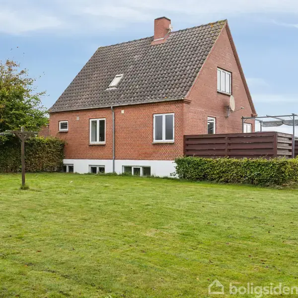 Et rødt murstenshus med skråt tag står i en grøn have med velplejet græsplæne. Til venstre en tørresnor, til højre en terrasse med et brunt hegn.