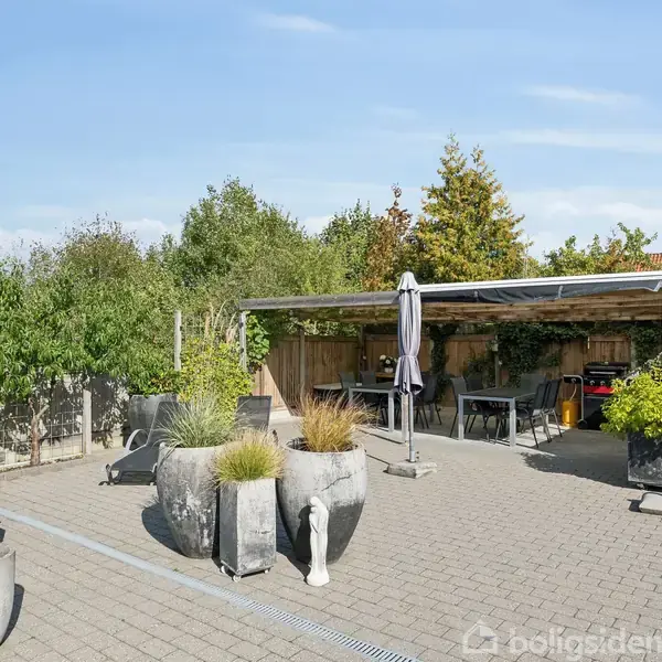 Terrasse med grå fliser og store, moderne plantekrukker. Overdækket spiseområde med bord, stole og en parasol. Træhegn og grønne træer omkranser området i solskinsvejr.