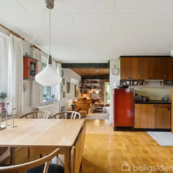Et spisebord står i en spisestue med moderne lamper hængende over. Baggrund: en køkkenvæg med træskabe, og en stue med en gul sofa ses i forlængelse.