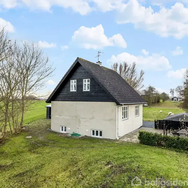 Et hvidt hus med sort tag står på en grøn græsplæne, omgivet af træer. Det er en klar dag med få skyer på himlen.