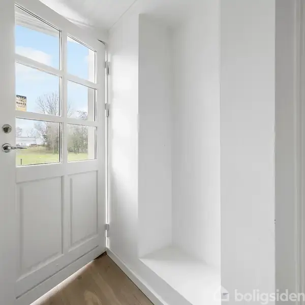 En hvid dør med glasruder står åben, og lys trænger ind. Døren fører ud til en græsplæne og træer i baggrunden. Indretningen er minimalistisk med lyse vægge.