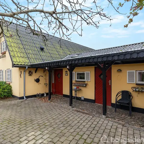 Et gulmalet hus med en grå, skrå tag, omgivet af flisebelagt gårdsplads. Enkelt facadeudsmykning med blomsterkasser ved vinduerne. Grå og blå himmel over det hele.