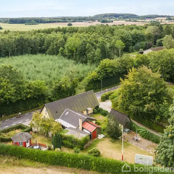 Et hus ligger omgivet af træer og buske i et landligt område. Der er en vej ved siden af huset, og marklandskab kan ses i baggrunden.