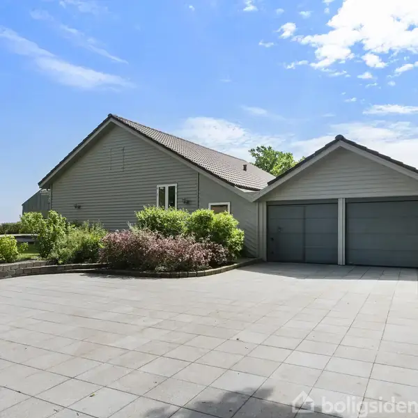 En gråmalet villa med to garageporte står stille omgivet af en flisebelagt forplads og velholdte planter, mens grønne træer og græsplæner indrammer ejendommen under en klar blå himmel.