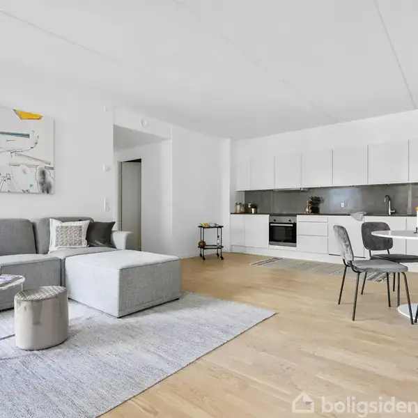 Grå sofa med puder står i et moderne åbent køkken-alrum. Et rundt bord med fire stole er placeret på trægulvet. Maleri og vase med blomster dekorerer rummet.