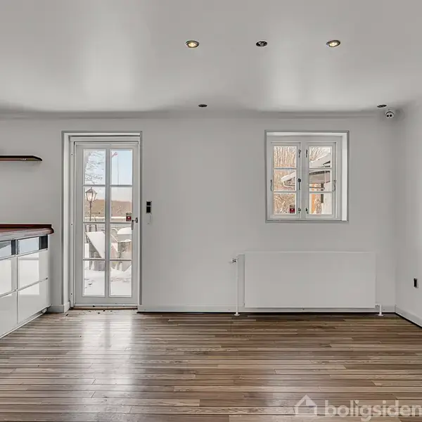 Et moderne køkken med lyst trægulv og hvid vægmaling. Køkkenøen har en vask og skabsenhed. Der er to vinduer og en glasdør, der leder til en terrasse.