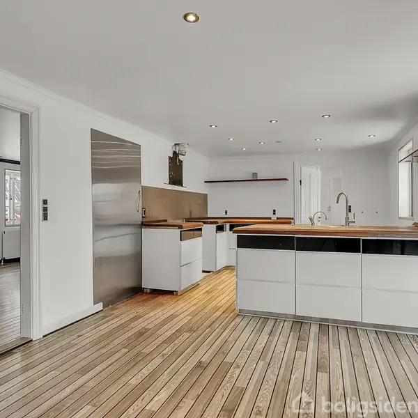 To store grantræer står i en snefyldt baghave foran et hus med skråtag, omgivet af andre huse og bevoksning, der skaber en fredelig vinterstemning.