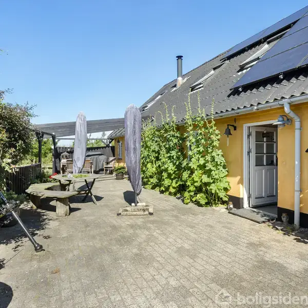 Rækkehus med solpaneler ovenpå, omgivet af planter klatrende op ad mur. To parasoller står ved siden af et udendørs siddeområde på fliser. Klar himmel i baggrunden.