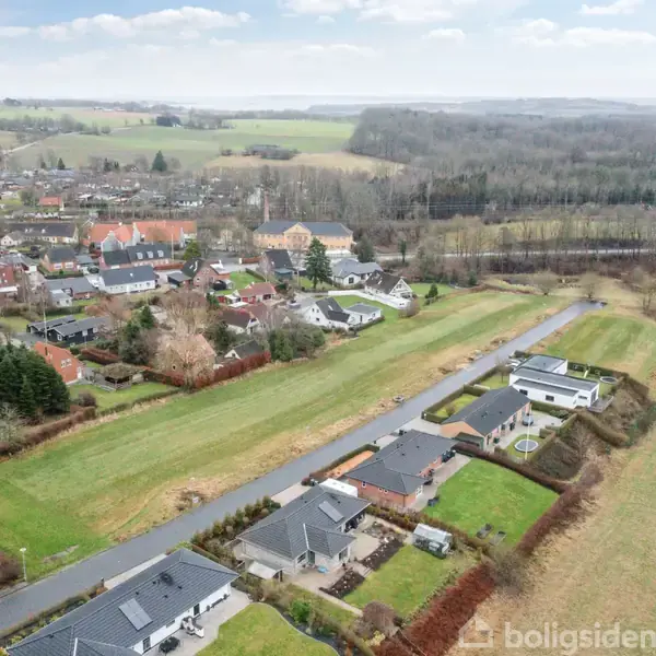 Byområde vist fra luften, med huse fordelt langs snoede veje, omgivet af åbne, grønne marker og skovområder i baggrunden under en let overskyet himmel.