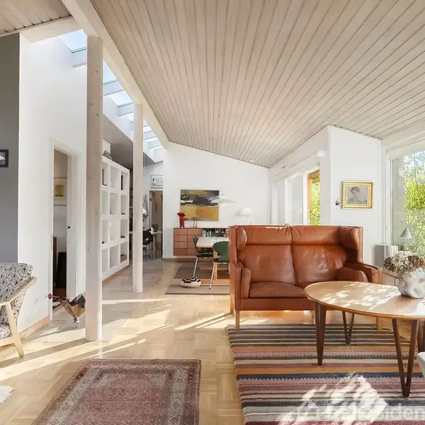 Brunt lædersofa står i en lys stue med skråloft og store vinduer, der giver udsigt til haven. Rummet er indrettet med stole, tæpper og kunst på væggene.