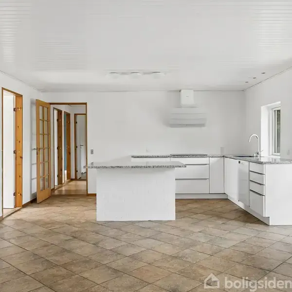 Køkken med hvide skabe og en central køkkenø, stående i et spartansk rum med flisegulve. Store vinduer og glasdør giver udsigt til det grønne udenfor.