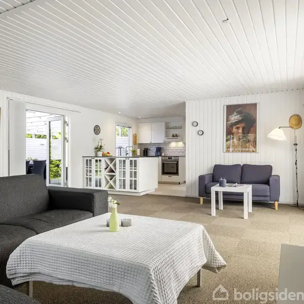 Stue med mørkegrå sofaer og hvid dug, åben forbindelse til køkken med hvidt interiør. Væggene har kunstværker, og gulvlampen tilføjer belysning. Store vinduer giver udsyn til have.